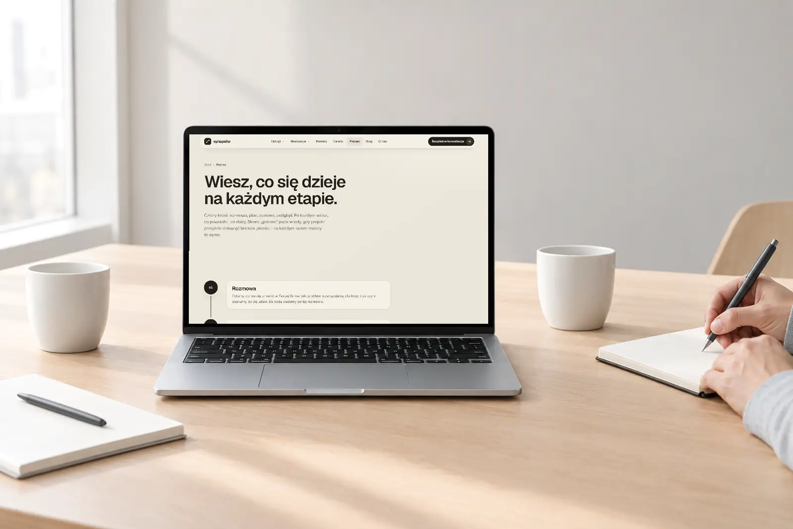 Laptop ze stroną procesu synapsite na stole do spotkań. Obok kubki i osoba robiąca notatki.